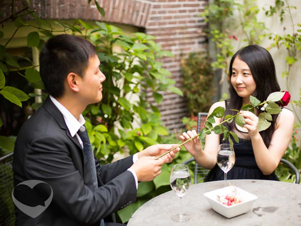 一位男士和一位女士相邻而坐，男士双手奉上一只玫瑰花递给女士，双方注视着彼此幸福的微笑。