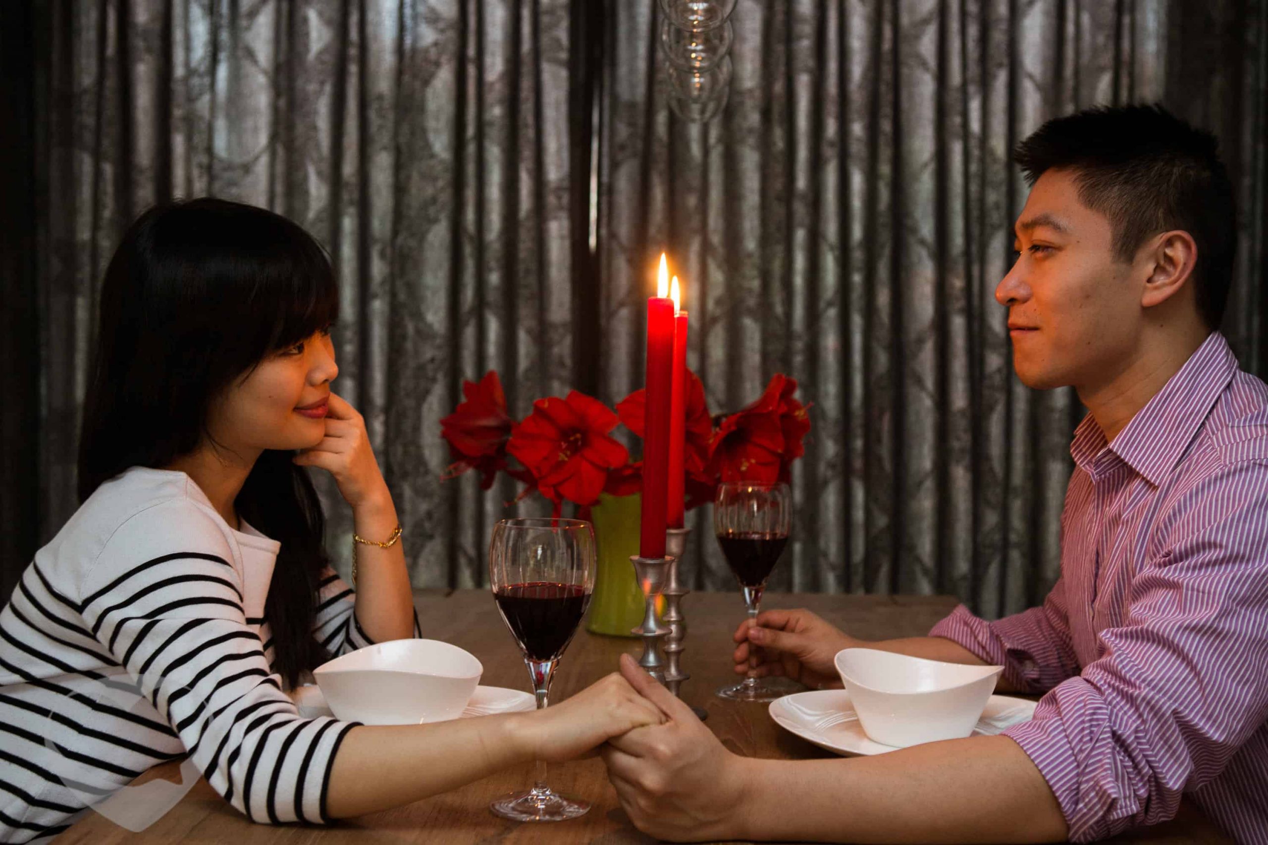 一位男士和一位女士面对面而座共进烛光晚餐，男士右手握着酒杯，左手牵着女士的手，女士的左手托着脸颊，双方注视着彼此幸福的微笑。
